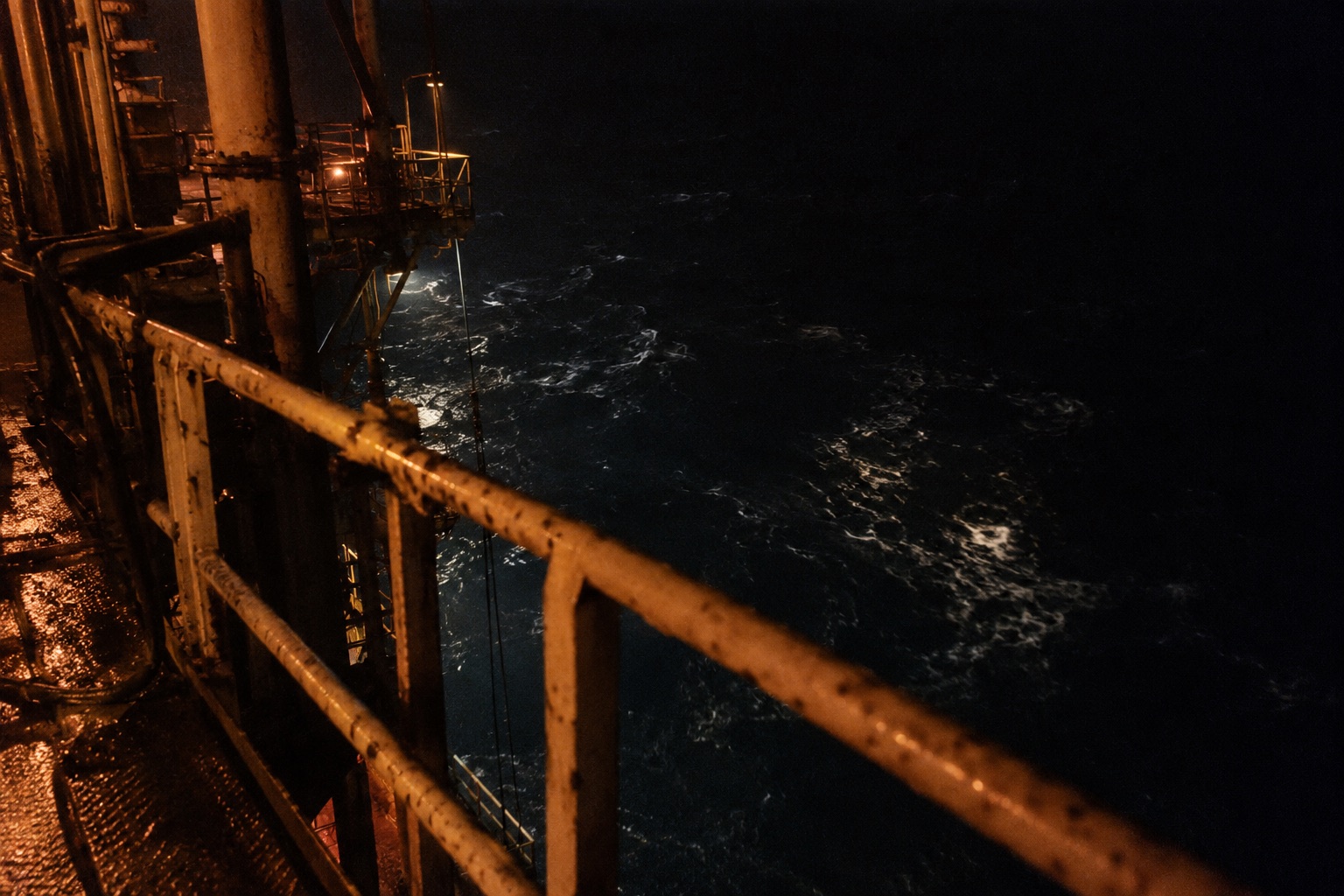 Night on the deck of an oil platform: a steel guard rail in the foreground, dark water far below catching faint light, and a warm glow from somewhere off-frame.